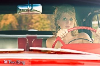 Bride in a birdcage veil behind the wheel of a red vintage car
