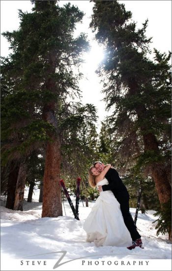 Bride and groom hug under pine tree with skis in the background