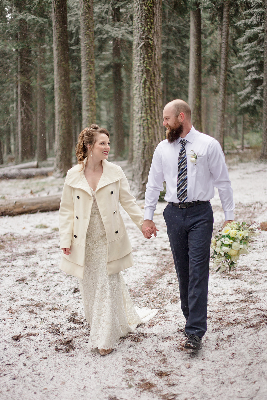 bride in lace dress wearing a white peacoat holding grooms hand in the mountains