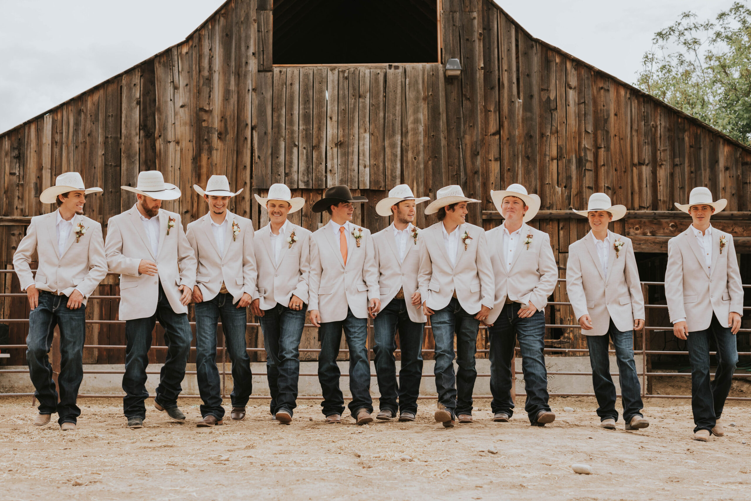 groom and groomsmen in front of barn with light colored jackets, jeans and cowboy hats
